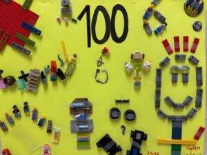 A bright yellow poster displays 100 in large black numbers, surrounded by various LEGO pieces and minifigures creatively arranged into patterns and shapes, with a few names and a date written at the bottom.