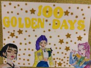 A white poster with 100 GOLDEN DAYS in large yellow letters, surrounded by glittery gold stars. At the bottom, there are colorful drawings of three stylish girls with different hair colors and fashion.