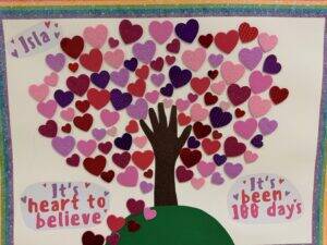 A handprint forms the trunk of a tree with branches covered in red, pink, and purple hearts. Speech bubbles read Isla, Its heart to believe, and Its been 100 days on a glittery rainbow-bordered background.