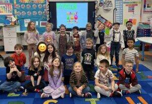 A group of young children pose and smile in a colorful classroom. Behind them, a screen displays Welcome! Happy 100 Days of School. The students are wearing festive clothing and some are dressed up as elderly people.